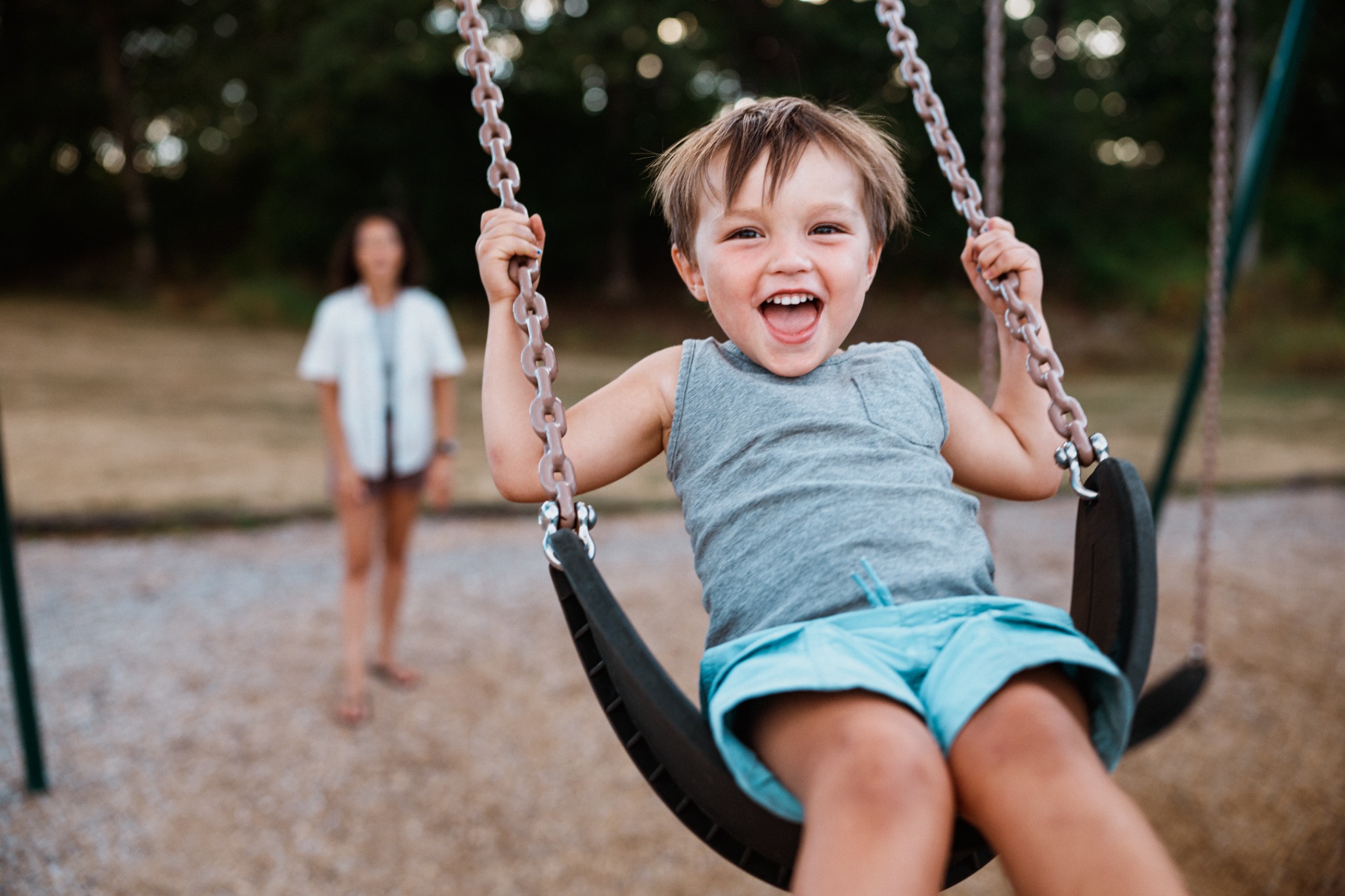 Young child smiling on a playground swing with a parent nearby