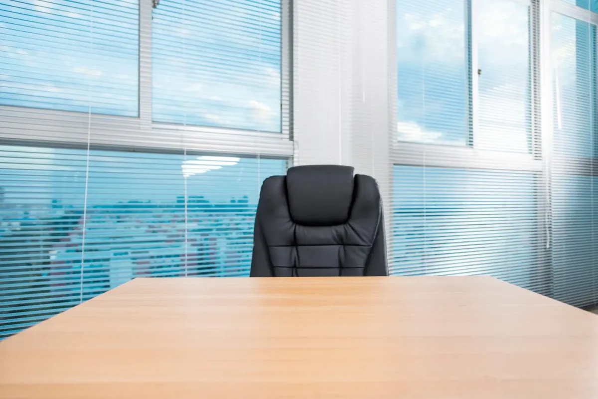 An empty executive chair at a boardroom table by a window — the succession question made visible