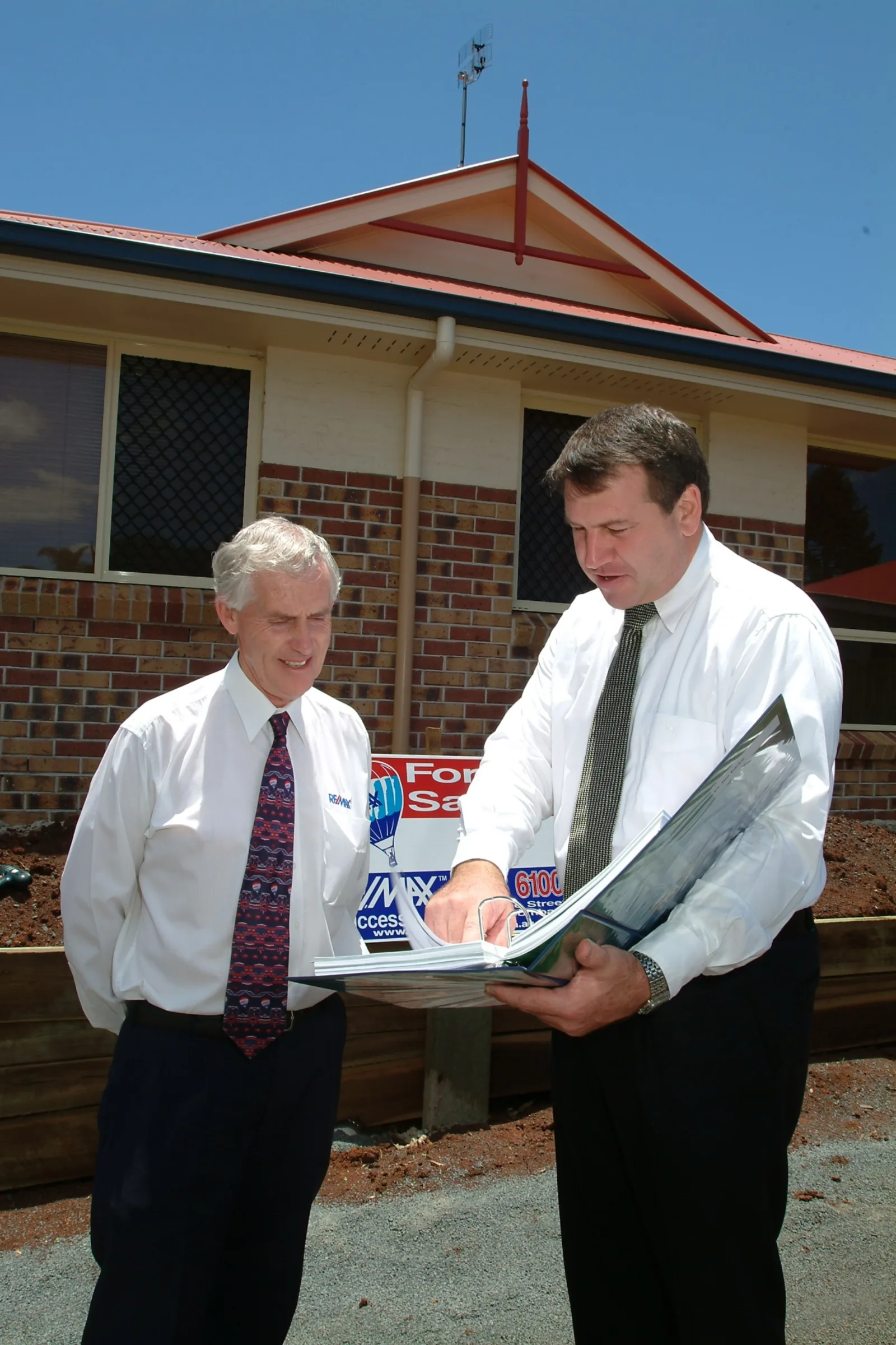 Two professionals reviewing a property listing portfolio in front of a residential home with a 'For Sale' sign