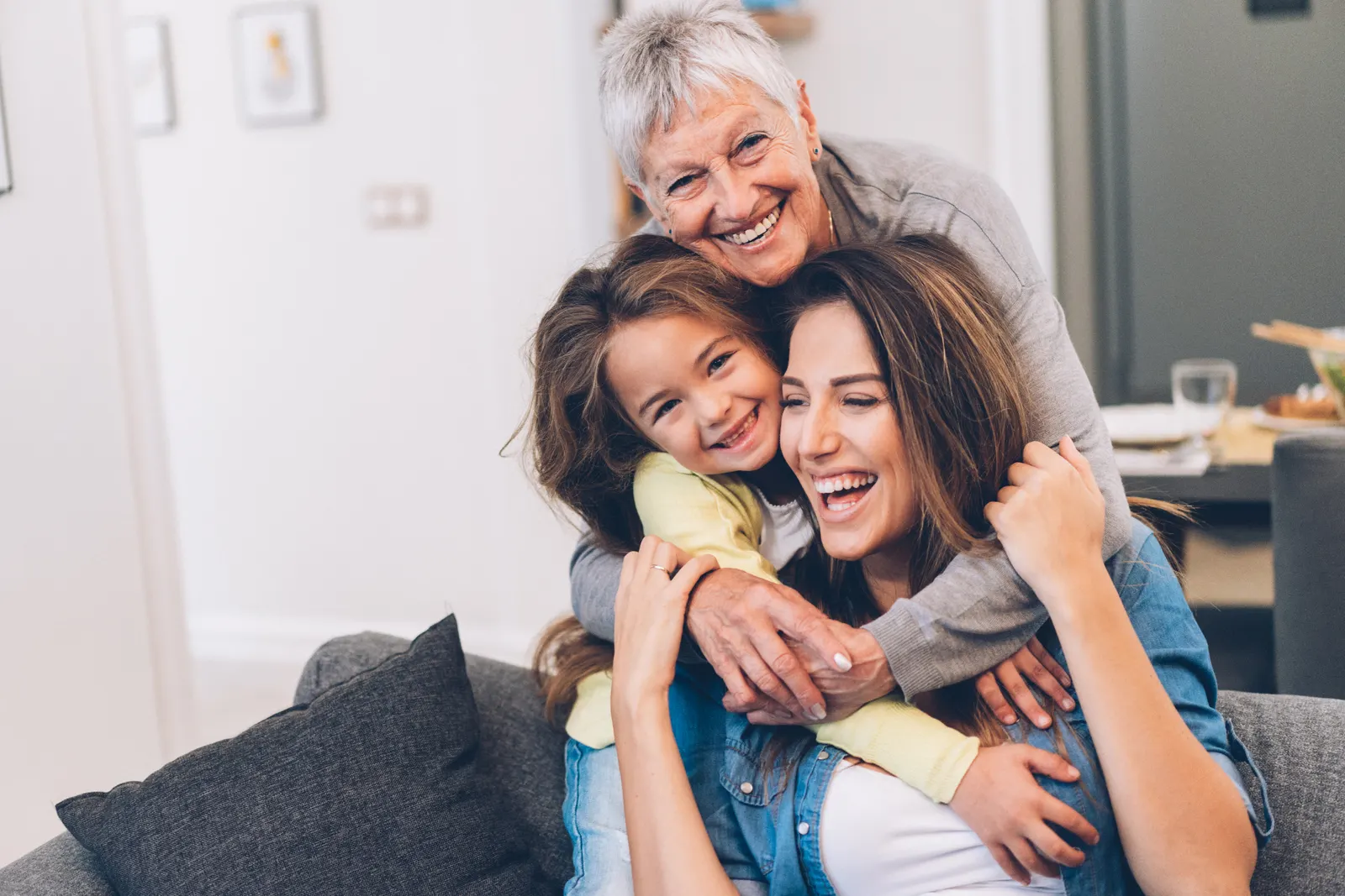 Three generations of a family — grandmother, mother and young daughter — laughing together at home.